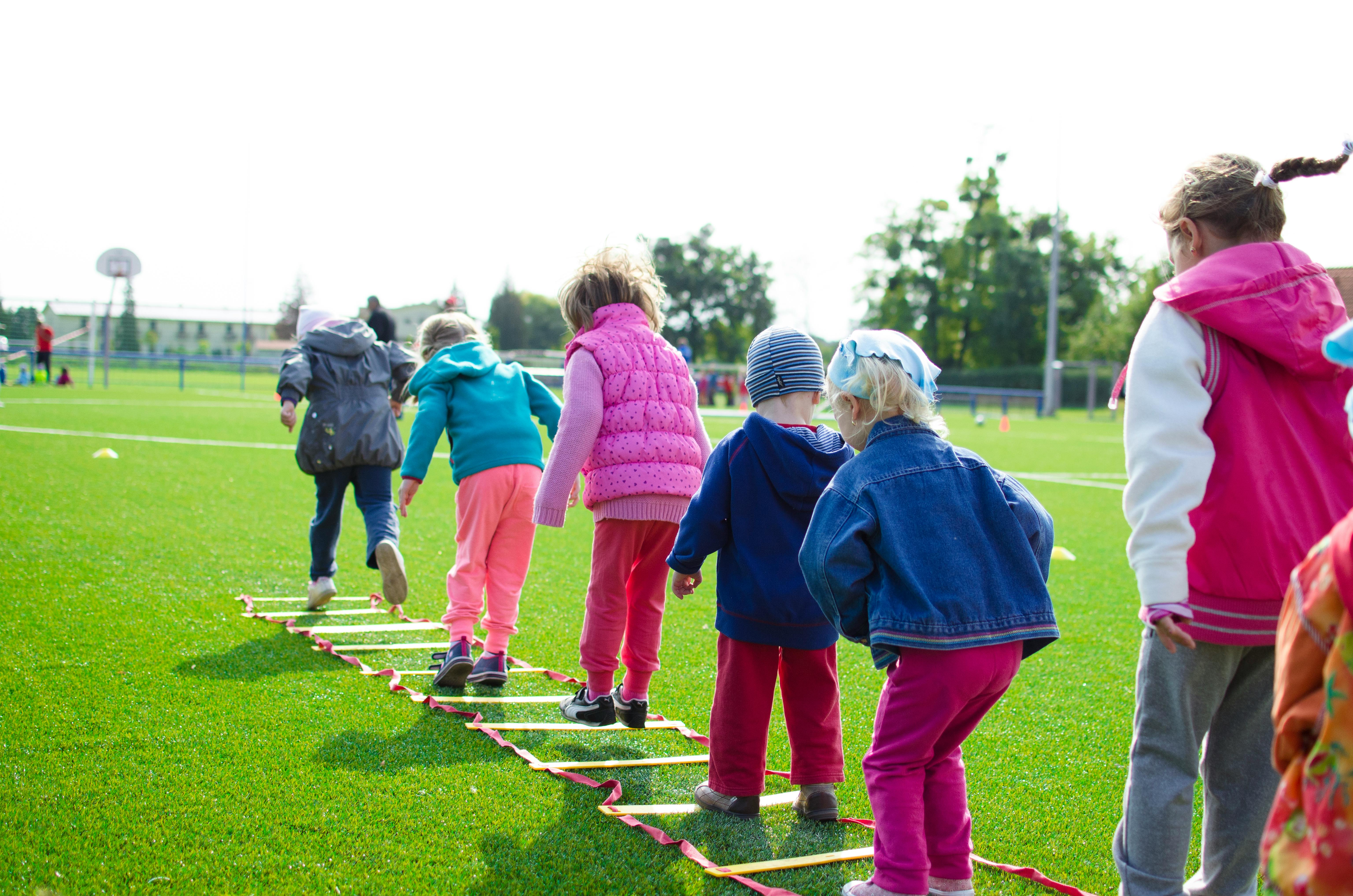 A group of young children participating in an outdoor activity on a grassy field. They are dressed in colorful jackets and pants, stepping carefully through a ladder laid flat on the ground as part of an exercise or game. Trees and playground equipment are visible in the background on a sunny day.