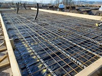 A section of concrete bounded by wood, a lattice of rebar covers the surface and several tubes emerge vertically from it.