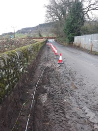 A dirt area next to a stone wall, separated from a surfaced road by cones.