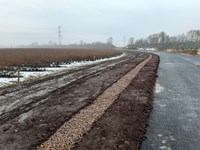 A dirt section of ground between a field and a road. There is a line of light brown gravel alongside the road.