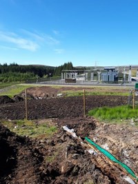 A trench reaching a dirt area, beyond the dirt area a substation is visible.