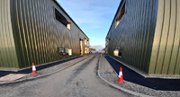 A road between two green-walled buildings, newly laid dark surfaced footpaths are present between the road and the buildings.