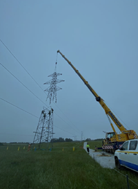 Contractors in PPE atop the base section of a metal transmission tower being dismantled. The upper section of the tower is suspeneded from a crane arm above.