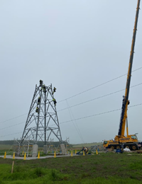 Contractors in PPE atop the base section of a metal transmission tower under construction.