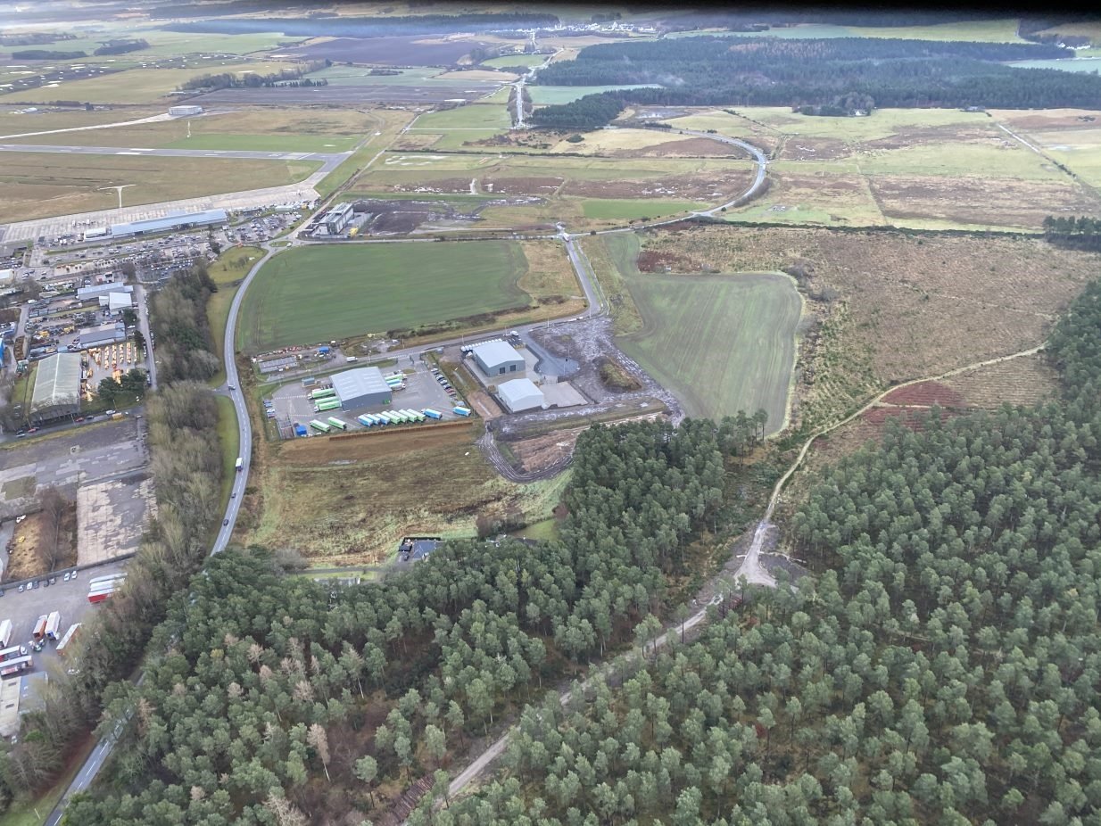 An aerial view of warehouses on an industrial development, surrounded on three sides by fields and a wooded area.