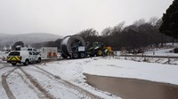 A large steel cylinder with cables wrapped around it, transported on a flat-bed trailer attached to a tractor, snow is falling in the air and present on the ground.