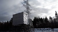 A covered scaffolding structure next to a metal transmission tower. Snow is present on the ground nearby.