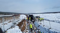 An excavated trench with repeating sets of metal structures along it, several thick black cables are being spooled out from a large drum at the far end of the trench by contractors in PPE. Snow covers the nearby ground.