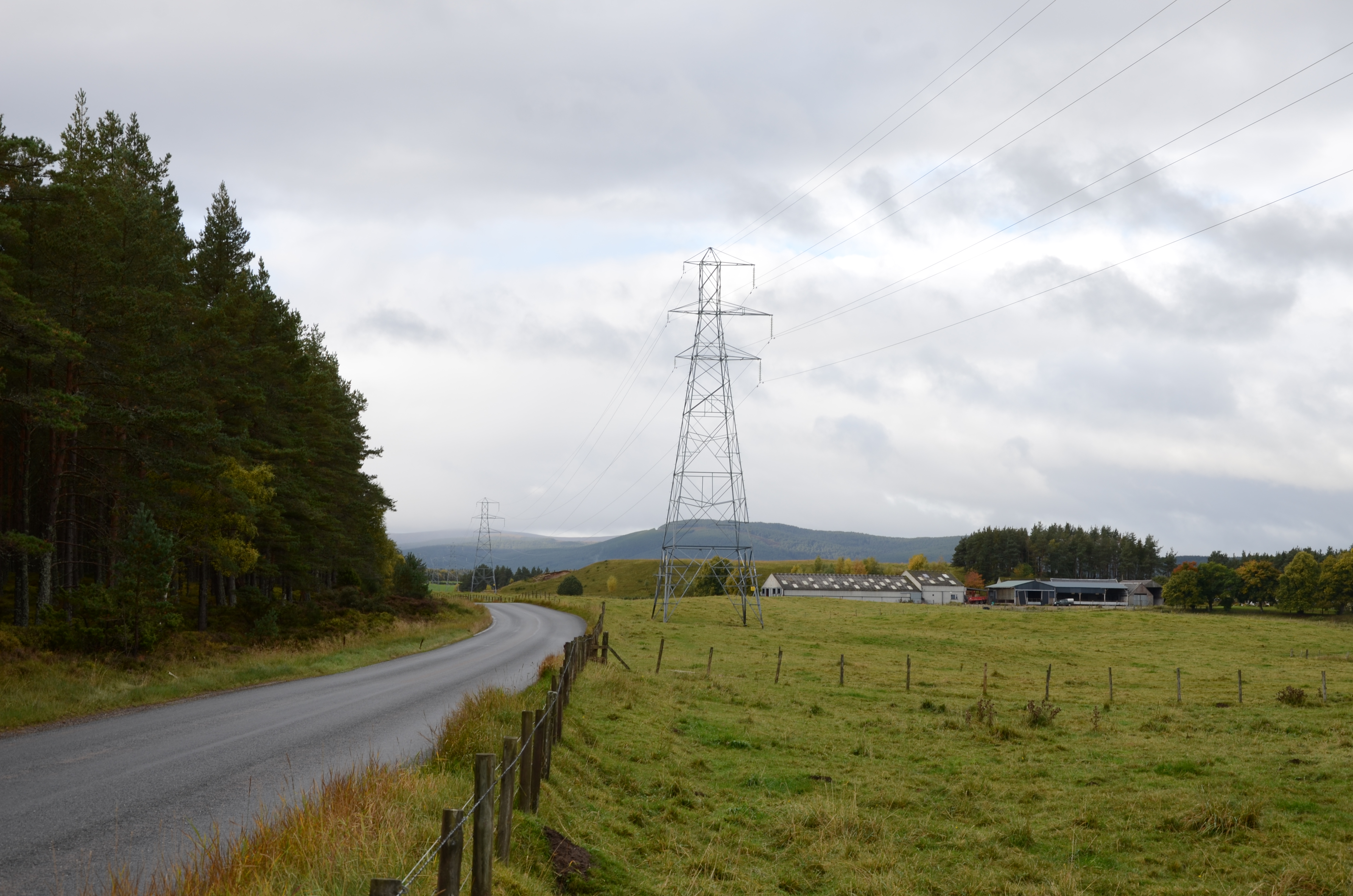 Metal transmission towers forming an overhead line over fields next to a road.
