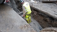 A contractor stands in a trench as material is poured into it in front of them.