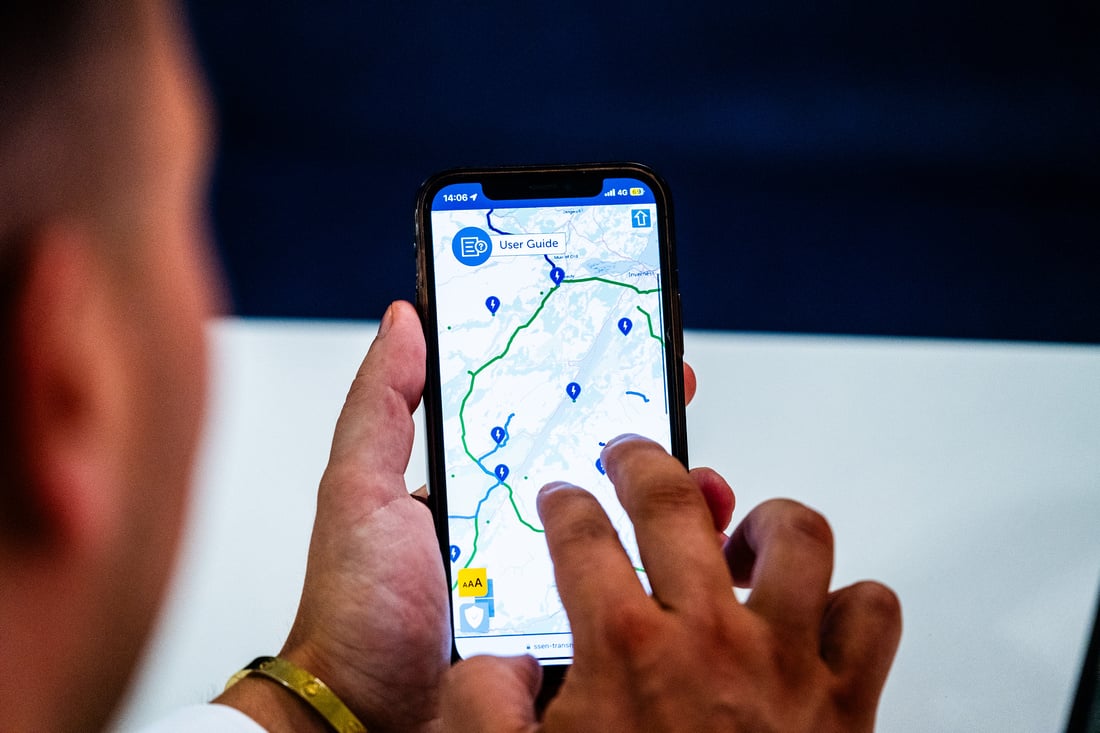 a man viewed from over the shoulder, focusing on his hands holding a smartphone. On the phone screen, a navigation app is open, displaying a map with a route and various location markers. The man is interacting with the map, possibly zooming in or selecting a destination.
