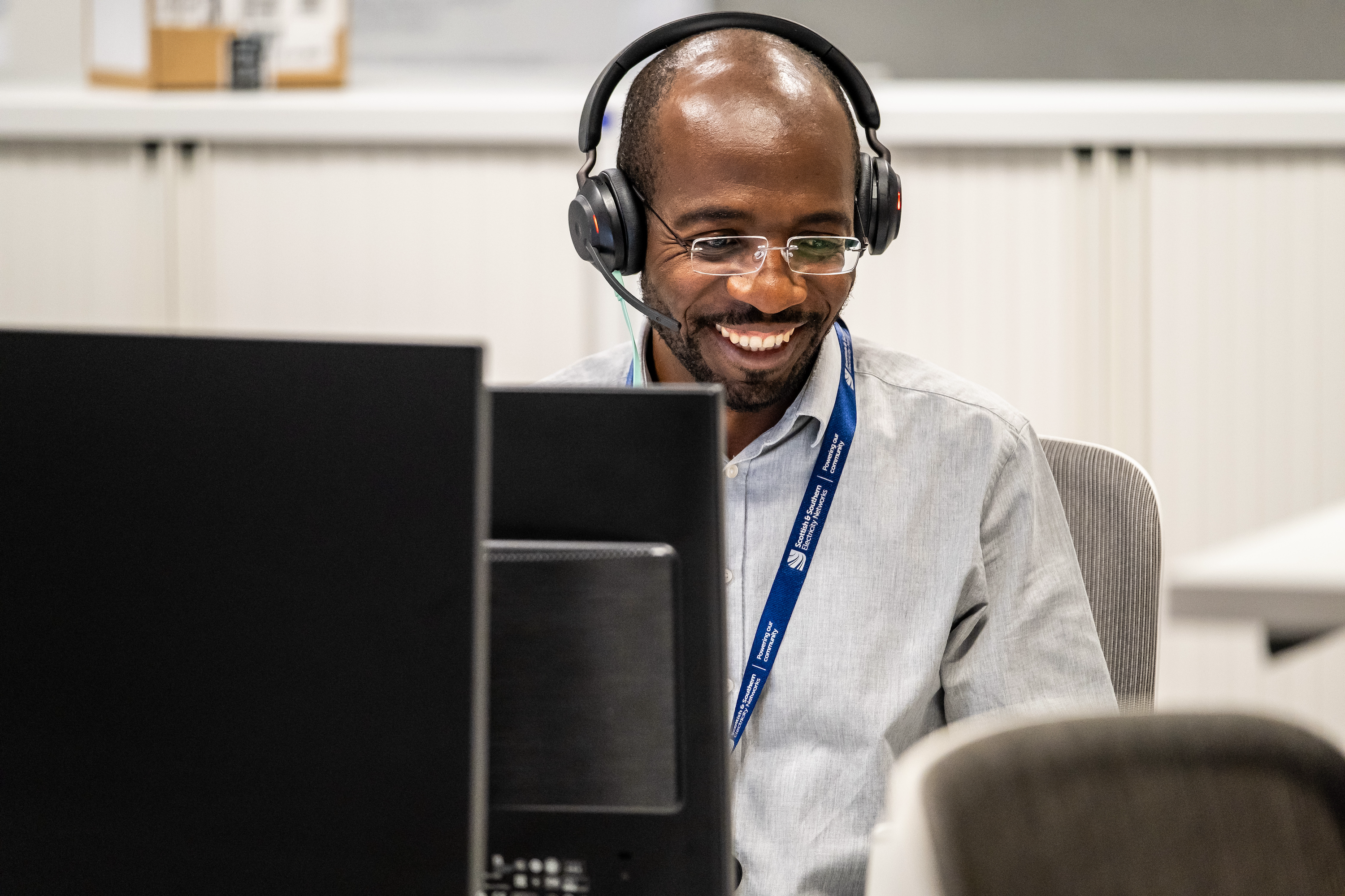 The image features a man working in a modern office setting. He is wearing a light grey shirt, glasses, and a headset with a microphone, suggesting he is engaged in a virtual call. The man is smiling broadly, looking at a computer screen, indicating he is having a pleasant interaction.